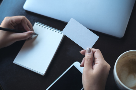 Top view image of a woman's hands holding empty white business card and writing on blank notebook with laptop and mobile phone on the tableの写真素材
