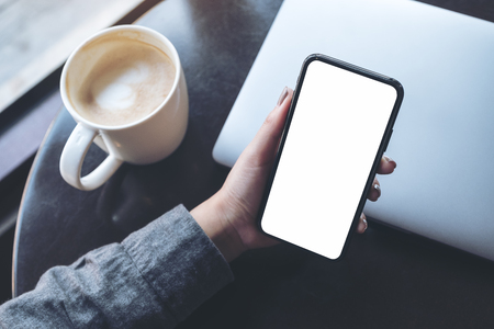 Mockup image of a woman's hand holding black mobile phone with blank white desktop screen with laptop and coffee cup on table in cafeの写真素材