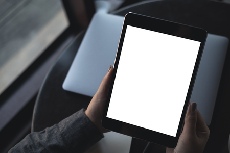 Top view mockup image of woman's hands holding black tablet pc with blank white desktop screen with laptop on table in cafeの写真素材