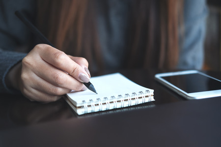 Closeup image of a woman's hands writing on notebook with mobile phone on the tableの写真素材