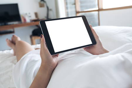 Mockup image of woman's hands holding black tablet pc with blank desktop white screen while lying in a white bedの写真素材