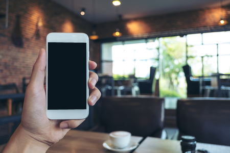 Mockup image of hand holding white mobile phone with blank black screen in modern cafeの写真素材