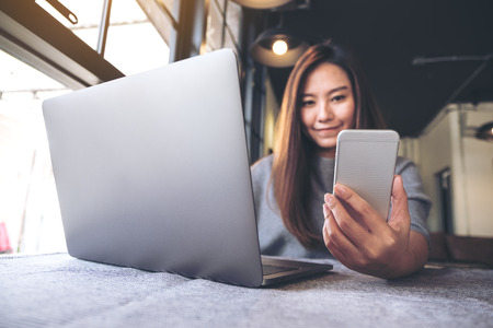 Closeup image of a beautiful Asian woman holding and looking at mobile phone while using laptop in cafeの写真素材