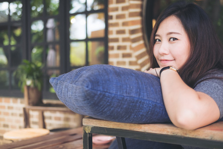A beautiful Asian woman sit with chin resting on her hands above a blue pillow with feeling happy and relax in cafe , bricks wall and nature backgroundの写真素材
