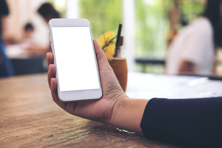Mockup image of a woman's hand holding white mobile phone with blank screen with a glass of iced coffee on wooden table in cafeの写真素材