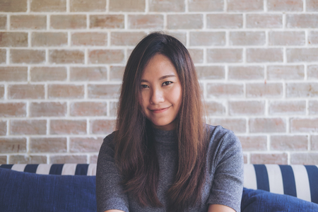 Closeup portrait image of a beautiful Asian woman with smiley face sitting on sofa with feeling good and relax with bricks wall backgroundの写真素材