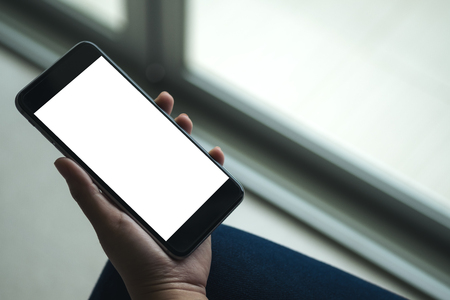 Mockup image of woman's hand holding black mobile phone with blank white screen on thigh with white tile floor and sliding door backgroundの写真素材