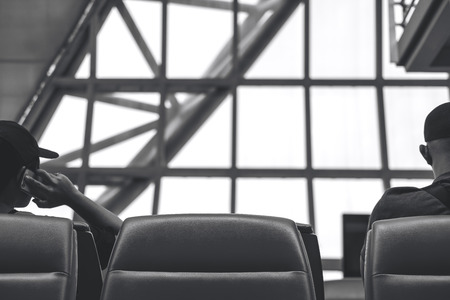 Black and white behind image of a man sitting and waiting for depart while another man talking on a phone in airportの写真素材