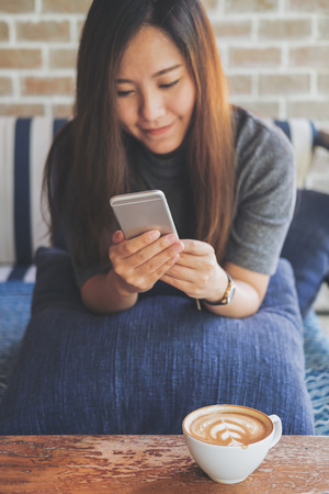 A beautiful Asian woman with smiley face using and looking at smart phone sitting on sofa with white latte coffee cup on wooden table in modern cafeの写真素材