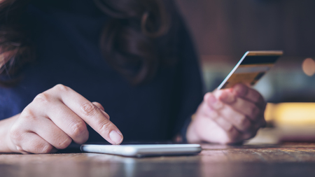 Closeup image of a woman's hand holding credit card and pressing at mobile phone on wooden table in officeの写真素材