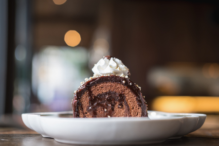 A piece of chocolate roll cake and whipped cream on the top in white ceramics plate on wooden table with blur background in cafeの写真素材