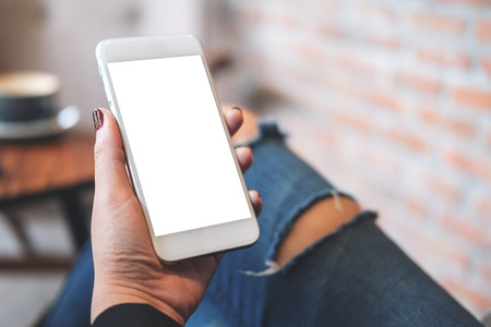 Mockup image of a woman's hand holding white mobile phone with blank  desktop screen on thigh in cafeの写真素材