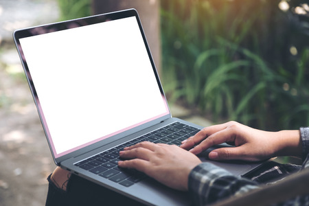 Mockup image of hands using and typing on laptop with blank white desktop screen , sitting outdoor on wooden chair and green nature backgroundの写真素材