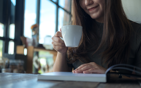 Closeup image of a beautiful Asian woman reading a book while drinking hot coffee in modern cafeの写真素材