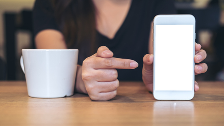 Mockup image of a beautiful woman holding , showing and pointing at white mobile phone with blank black screen with white coffee cup on wooden table in restaurantの写真素材