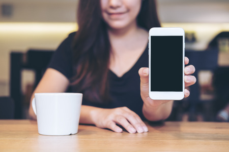 Mockup image of an Asian beautiful woman holding and showing white mobile phone with blank black screen and white coffee coupon wooden table in cafeの写真素材