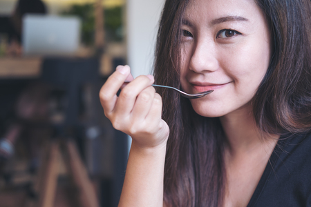 Closeup image of an Asian woman with smiley face eating food by spoon in modern cafeの写真素材