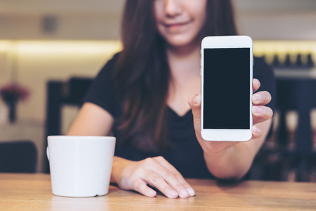 Mockup image of an Asian beautiful woman holding and showing white mobile phone with blank black screen and white coffee coupon wooden table in cafeの写真素材