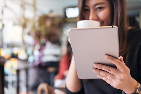 Closeup image of a beautiful Asian woman with smiley face holding and using tablet pc while drinking coffee in modern cafeの写真素材