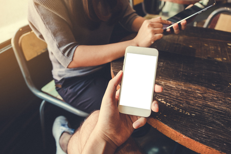 Mockup image of a hand holding white mobile phone with blank desktop screen in vintage cafe with a woman using tablet in backgroundの写真素材