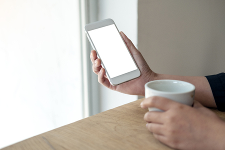 Mockup image of hands holding white mobile phone with blank screen with a cup of coffee on wooden table in cafeの写真素材