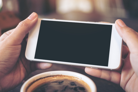 Mockup image of hands holding white mobile phone with blank black screen for watching and playing games with a cup of coffee on wooden table in cafeの写真素材