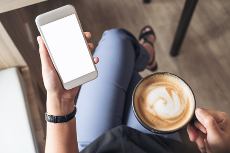 Top view mockup image of a woman's hand holding white mobile phone with blank desktop screen while drinking coffee in cafeの写真素材