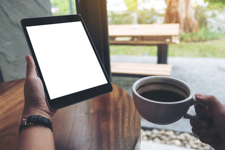 Mock up image of hands holding black tablet pc with white blank screen while drinking coffee in cafeの写真素材