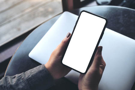 Mockup image of a woman's hand holding black mobile phone with blank white desktop screen with laptop on table in cafeの写真素材