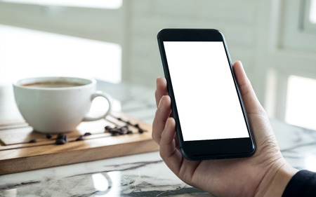 Mockup image of hands holding black mobile phone with blank white screen with a cup of coffee in cafeの写真素材