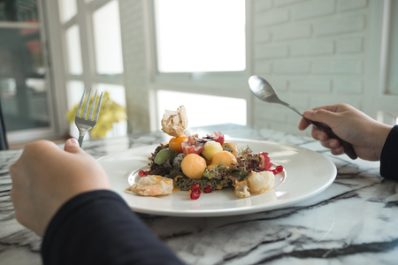 A woman holding spoon and fork and preparing to eat mixed fruits salad with fried shrimps on the table in restaurantの写真素材