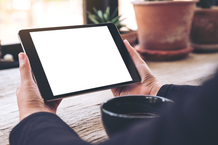Mockup image of hands holding black tablet pc with blank white screen and coffee cup on vintage...