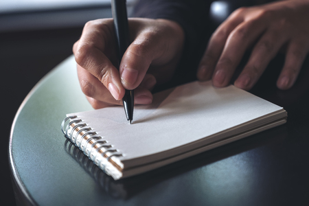 Closeup image of a woman's hands writing down on a white blank notebook on table の写真素材