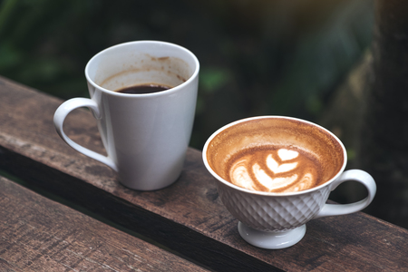 Closeup image of two cups of black coffee and latte with heart latte art on wooden bench in green nature backgroundの写真素材