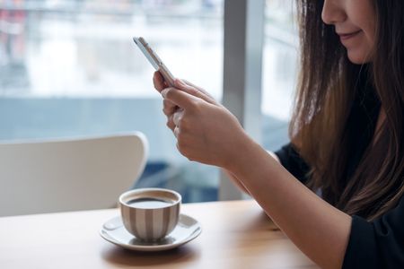 Closeup image of a beautiful Asian woman with smiley face holding and using smart phone with coffee cup on wooden table in cafeの写真素材