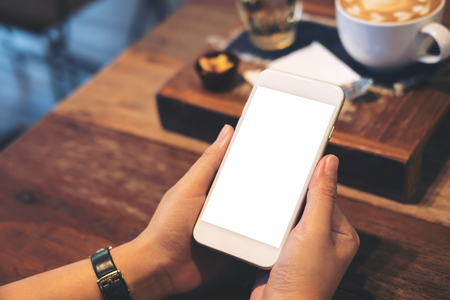 Mockup image of a woman's hands holding white mobile phone with blank screen with coffee cup on wooden table in vintage cafeの写真素材