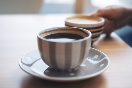 Closeup image of Americano coffee cup and woman's hand holding latte coffee cup on vintage wooden table in cafeの写真素材