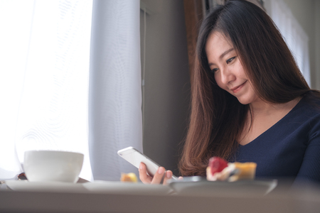 Closeup image of a smiley beautiful Asian woman holding , using and looking at smart phone with white coffee cup and dessert plate on wooden table in vintage cafeの写真素材