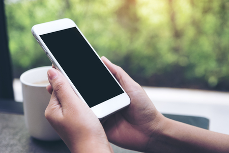 Mockup image of hands holding white mobile phone with blank black desktop screen with coffee cup on table in cafeの写真素材