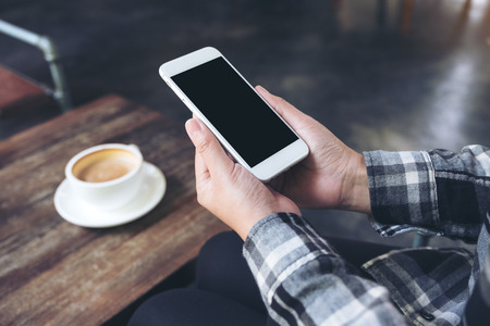 Mockup image of a woman's hands holding white mobile phone with blank black desktop screen with coffee cup on wooden table in cafeの写真素材