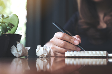 Closeup image of a businesswoman working and writing down on a white blank notebook with screwed up papers on tableの写真素材
