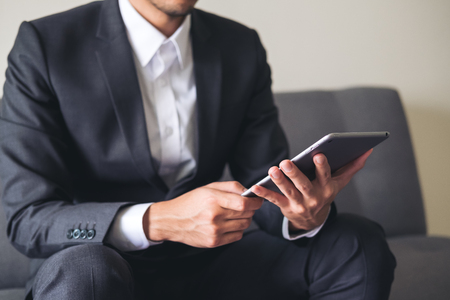 A business man in a gray suit sitting on sofa holding , using and looking at tablet pc for work in officeの写真素材