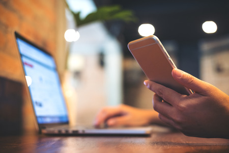 A business woman holding mobile phone while using laptop on wooden table in officeの写真素材