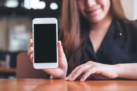 Mockup image of a beautiful woman holding and showing white mobile phone with blank black screen in restaurantの写真素材