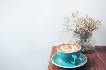 Copy space image of a blue cup of hot latte coffee and dry flowers in a vase on vintage wooden table in coffee shopの写真素材