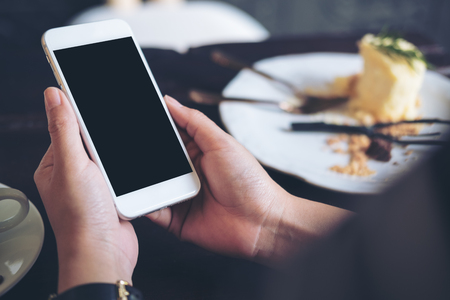 Mockup image of hands holding white mobile phone with blank black screen with a plate of cake on wooden table in restaurantの写真素材