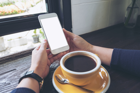 Mockup image of woman's hands holding white mobile phone with blank screen and yellow coffee cup on wooden table in cafeの写真素材