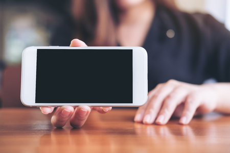 Mockup image of a woman holding and showing white horizontal mobile phone with blank black screen in restaurantの写真素材