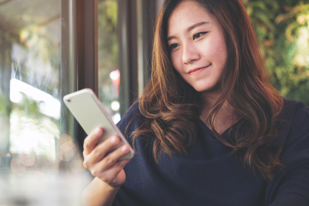 A beautiful Asian woman with smiley face using and looking at smart phone in modern green nature cafeの写真素材