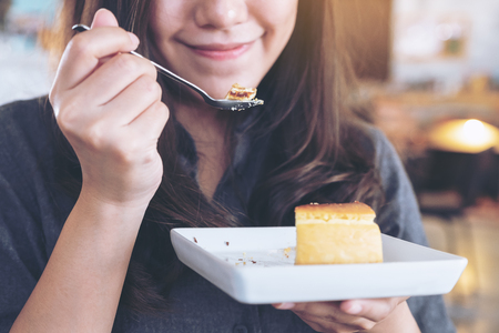 Closeup image of a happy woman holding and enjoy eating cheese cake with spoon in cafeの写真素材
